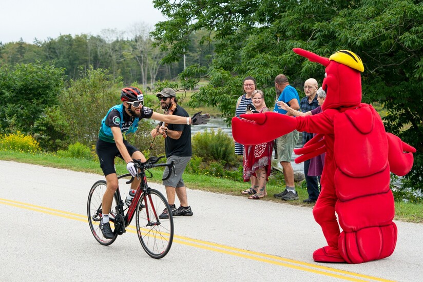 Une image d'un cycliste saluant une mascotte en forme de homard, avec des personnes encourageant le cycliste et un violoniste derrière lui.